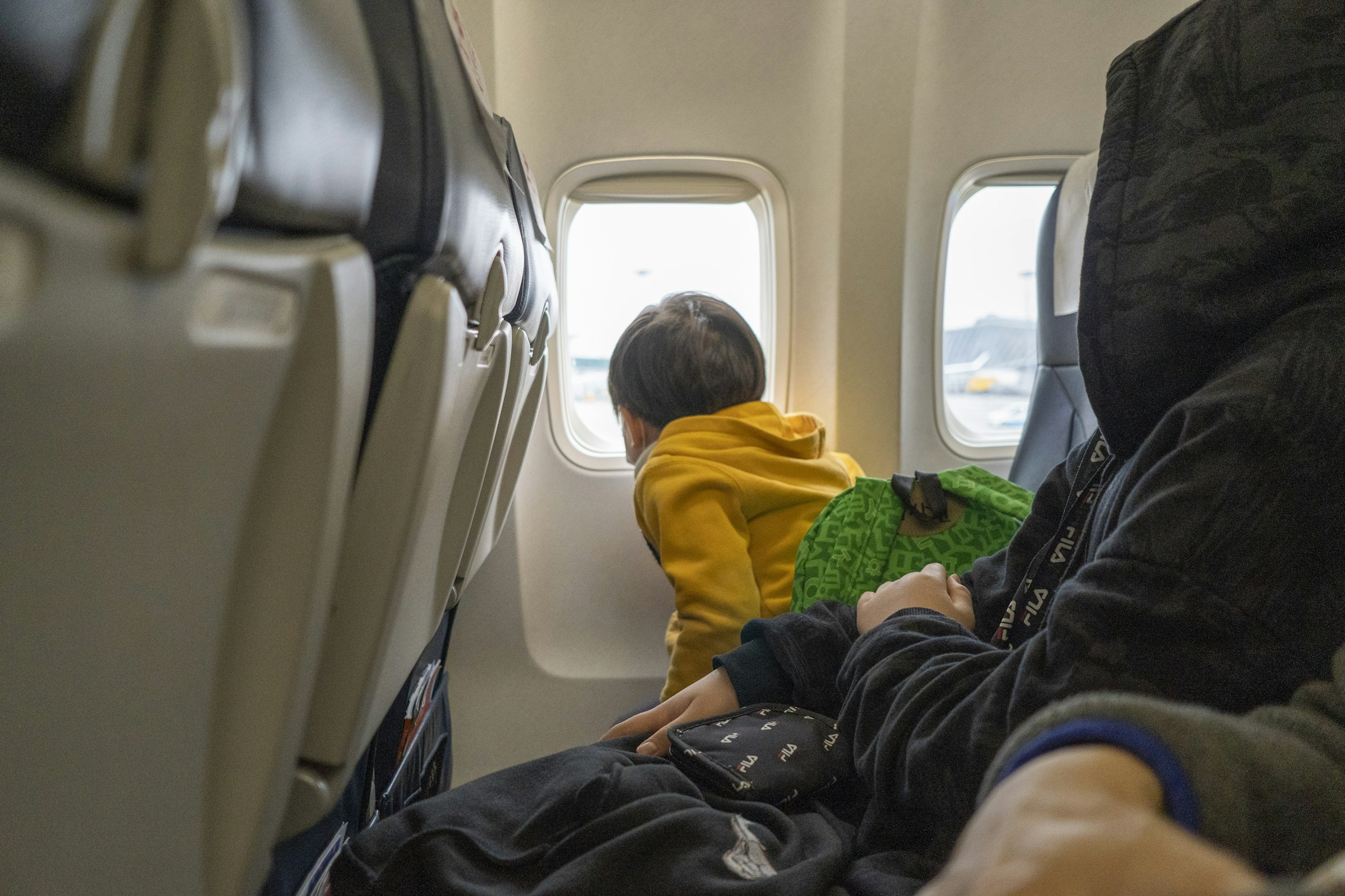 A small child in a yellow jacket kneeling on an airplane seat and looking out the window
